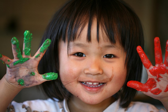 A child smiles while holding up her hands. One hand is covered in red paint and the other in green.