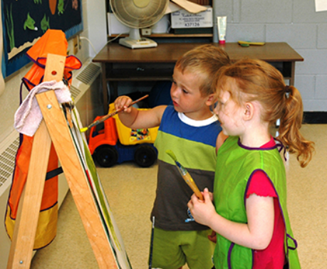 Two children stand in front of a wooden easel painting with large wooden paint brushes.