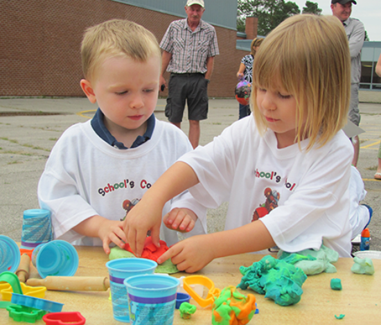 Two children in School's Cool T-shirts are playing with playdoh on a table outdoors.